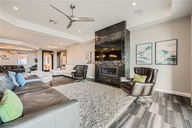 living area with visible vents, baseboards, a premium fireplace, ceiling fan with notable chandelier, and a raised ceiling