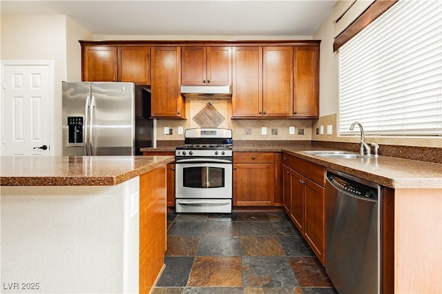 kitchen featuring stone finish floor, under cabinet range hood, a sink, stainless steel appliances, and brown cabinetry