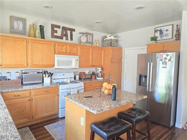 kitchen with a center island, dark wood-type flooring, a kitchen bar, light stone counters, and white appliances