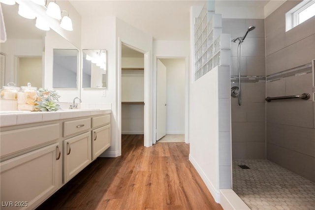 full bath featuring vanity, wood finished floors, baseboards, tiled shower, and a walk in closet