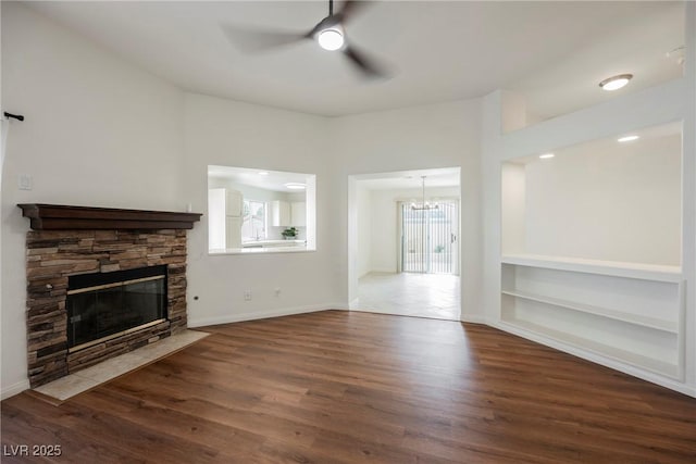 unfurnished living room featuring built in features, wood finished floors, baseboards, a stone fireplace, and ceiling fan with notable chandelier