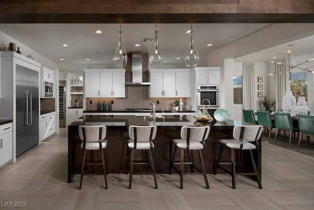 kitchen with dark countertops, built in appliances, a kitchen breakfast bar, and wall chimney exhaust hood