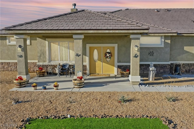 exterior entry at dusk featuring a tile roof, stone siding, and stucco siding