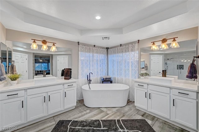 bathroom featuring a tray ceiling, a soaking tub, two vanities, a stall shower, and a sink