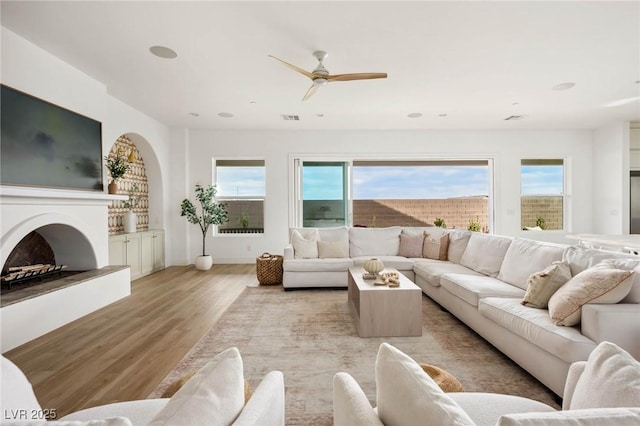 living area with visible vents, a fireplace with raised hearth, a ceiling fan, arched walkways, and light wood finished floors