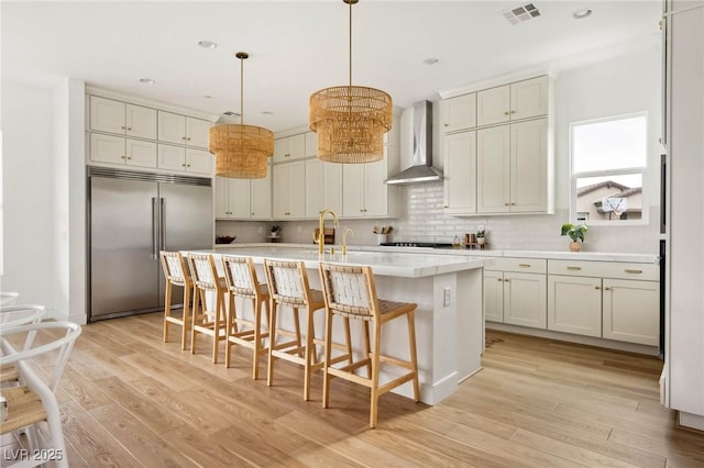 kitchen featuring light wood finished floors, visible vents, decorative backsplash, wall chimney exhaust hood, and built in fridge