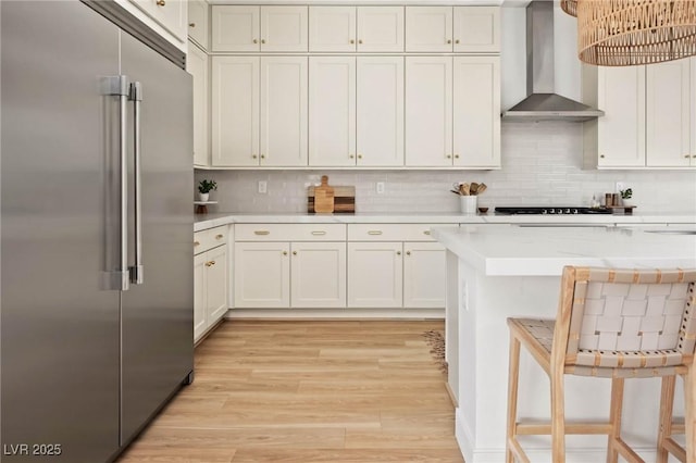 kitchen with backsplash, light countertops, high end refrigerator, light wood-style floors, and wall chimney exhaust hood