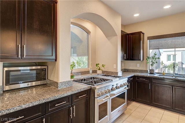 kitchen featuring a sink, dark brown cabinetry, appliances with stainless steel finishes, light tile patterned flooring, and stone counters