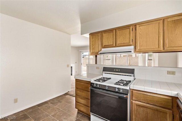 kitchen with baseboards, tile counters, under cabinet range hood, gas range, and brown cabinets