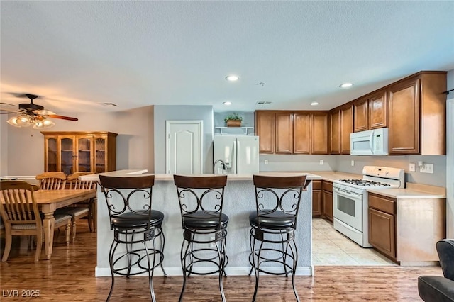 kitchen with white appliances, a ceiling fan, a kitchen island with sink, light countertops, and a kitchen breakfast bar