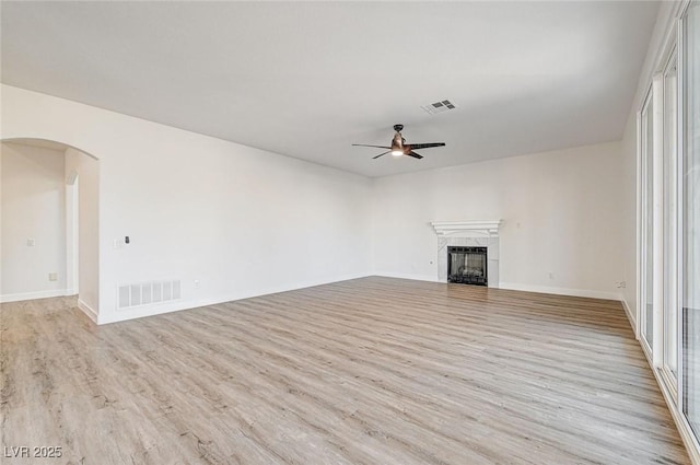 unfurnished living room with light wood finished floors, visible vents, ceiling fan, a tiled fireplace, and arched walkways