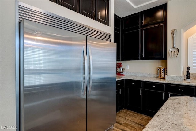 kitchen with light stone counters, wood finished floors, dark cabinetry, and built in fridge