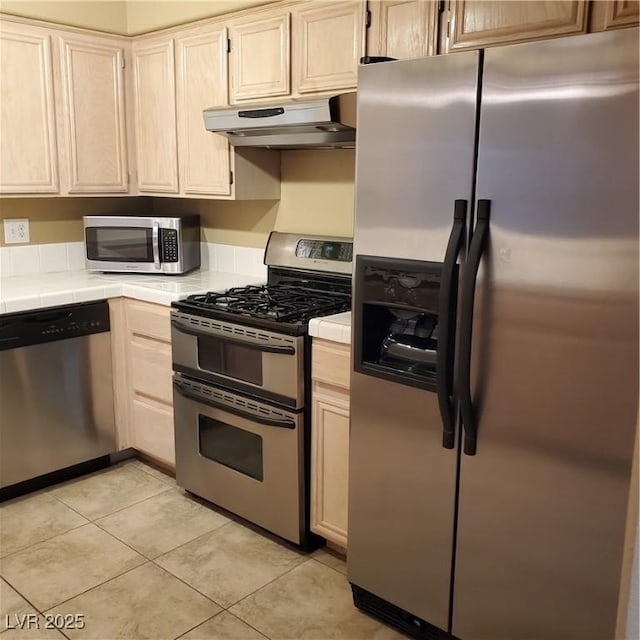 kitchen with light brown cabinets, tile counters, under cabinet range hood, light tile patterned floors, and stainless steel appliances