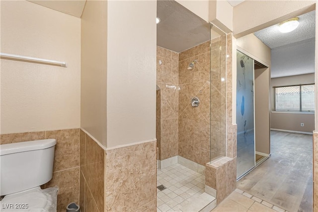 full bathroom featuring wainscoting, toilet, a textured ceiling, and tiled shower