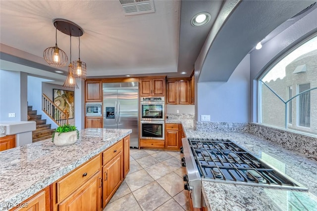 kitchen with visible vents, pendant lighting, brown cabinetry, built in appliances, and light stone countertops