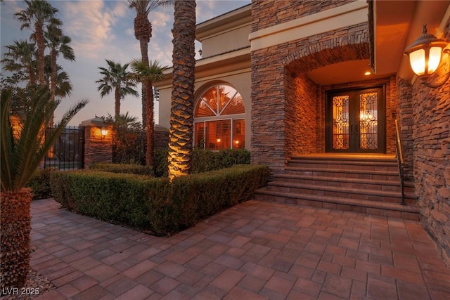 exterior entry at dusk featuring stone siding, stucco siding, french doors, and fence