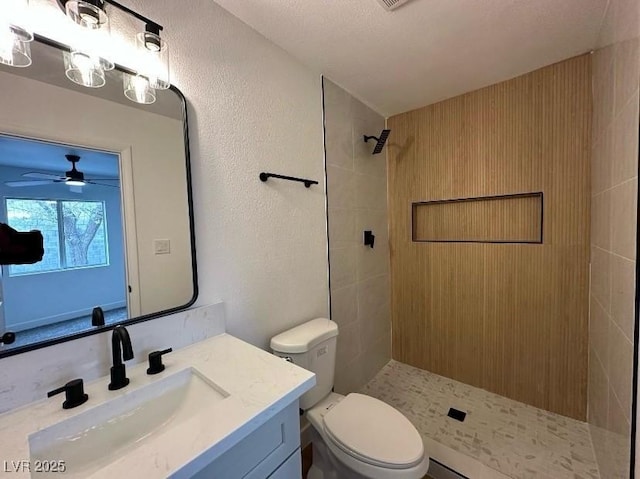 full bath featuring toilet, tiled shower, ceiling fan, vanity, and a textured wall