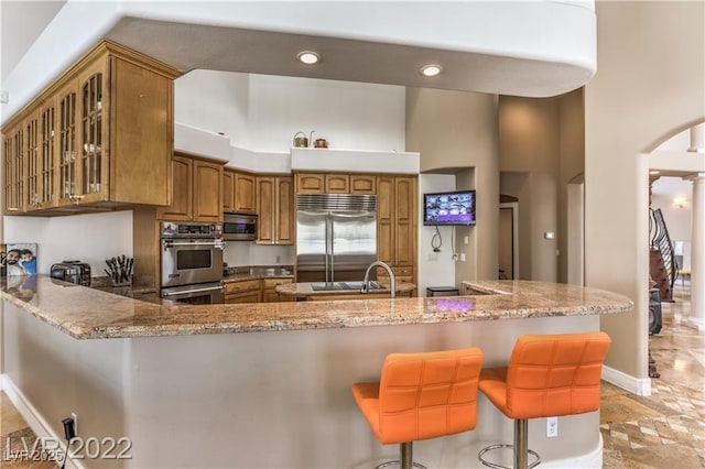 kitchen with light stone countertops, brown cabinetry, a peninsula, glass insert cabinets, and appliances with stainless steel finishes