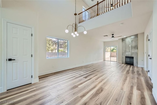 unfurnished living room with light wood-style flooring, ceiling fan with notable chandelier, a fireplace, baseboards, and a towering ceiling