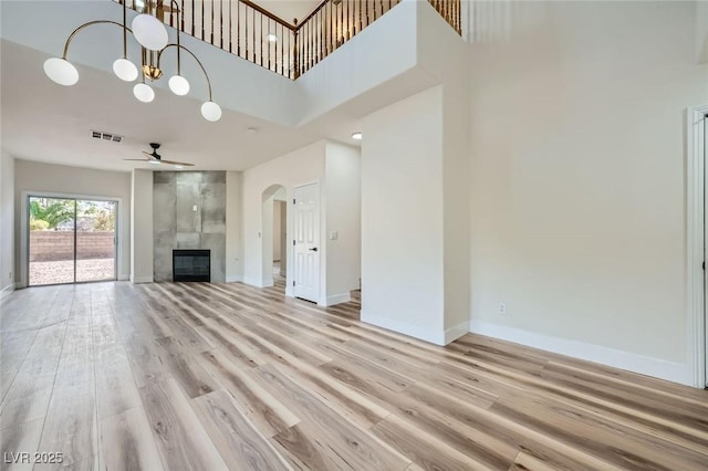 unfurnished living room with baseboards, light wood-type flooring, ceiling fan with notable chandelier, a tile fireplace, and arched walkways