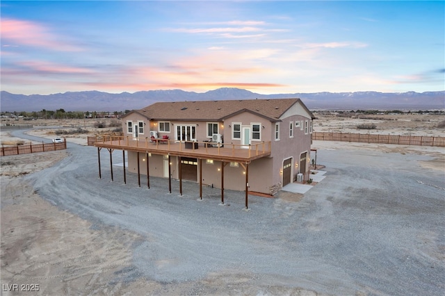 back of property at dusk featuring a deck with mountain view, fence, dirt driveway, and stucco siding