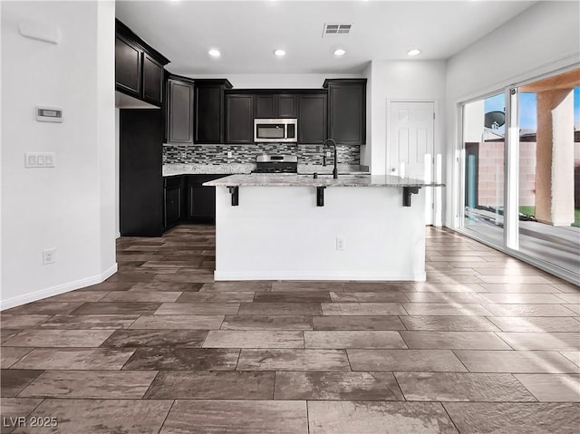 kitchen with light stone counters, a center island with sink, visible vents, stainless steel appliances, and backsplash