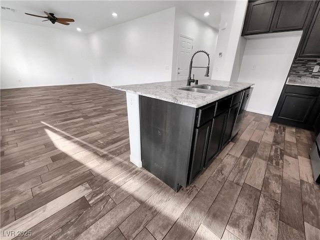 kitchen with a ceiling fan, light stone countertops, an island with sink, a sink, and dark wood-type flooring