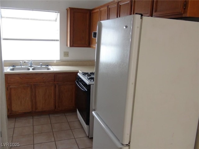 kitchen with a sink, freestanding refrigerator, brown cabinetry, light tile patterned floors, and gas range