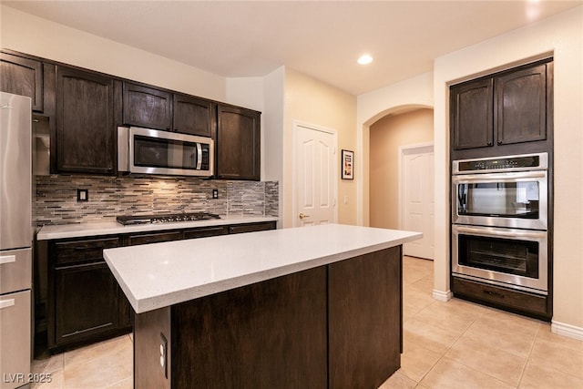 kitchen featuring light tile patterned flooring, arched walkways, dark brown cabinetry, appliances with stainless steel finishes, and backsplash