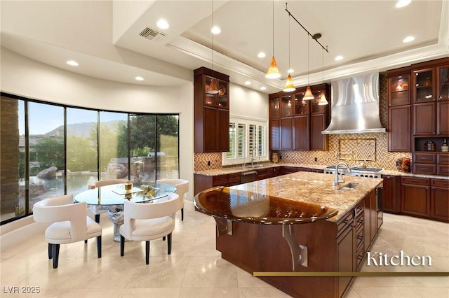 kitchen featuring visible vents, an island with sink, light stone counters, a raised ceiling, and wall chimney exhaust hood