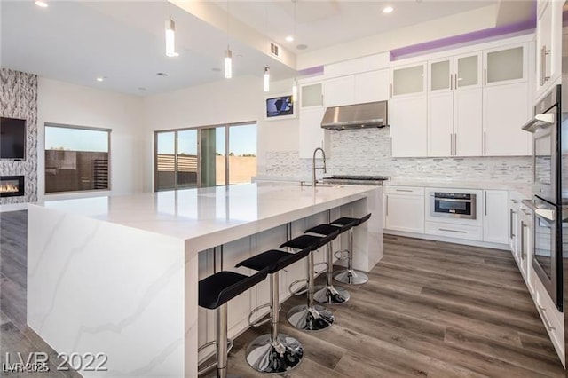 kitchen with dark wood-style flooring, decorative backsplash, white cabinets, under cabinet range hood, and a large fireplace