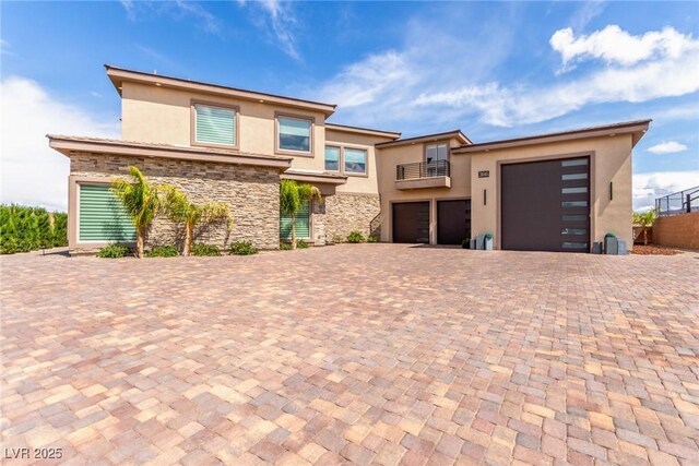 prairie-style home with stone siding, a balcony, decorative driveway, and stucco siding