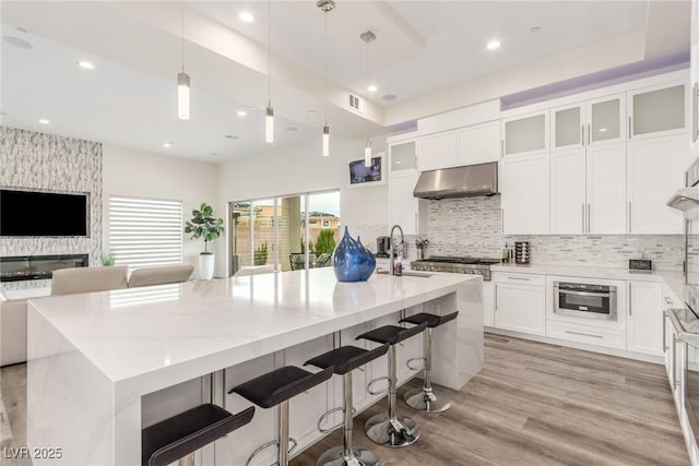 kitchen featuring visible vents, under cabinet range hood, a sink, light wood-style floors, and backsplash