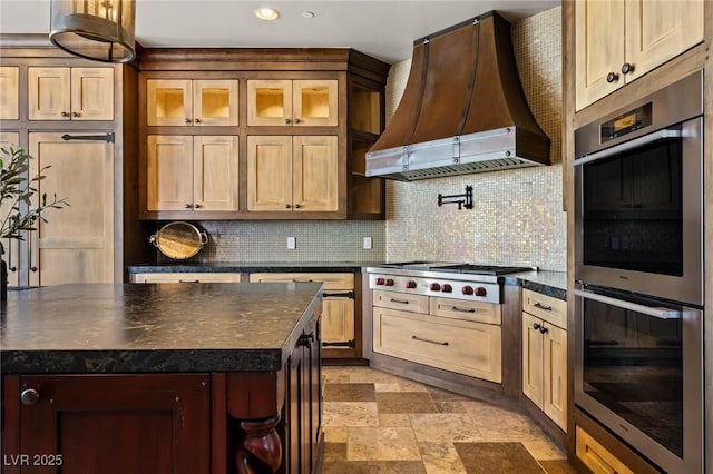 kitchen with custom exhaust hood, stone finish floor, appliances with stainless steel finishes, dark countertops, and tasteful backsplash