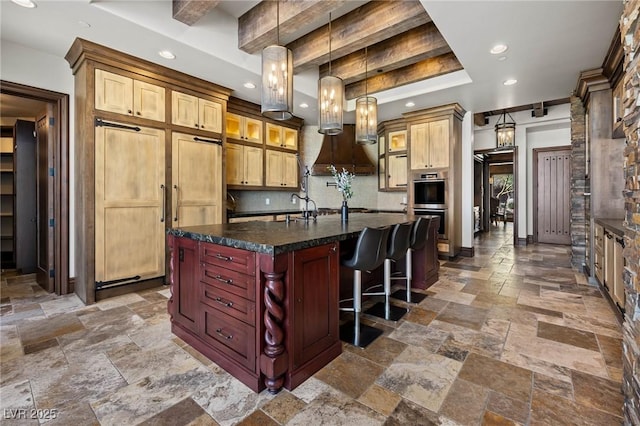 kitchen with tasteful backsplash, premium range hood, a center island with sink, stone tile floors, and stainless steel double oven