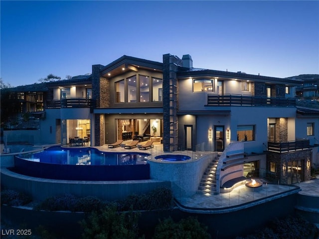 back of property at dusk with a balcony, an in ground hot tub, stucco siding, a patio area, and an infinity pool