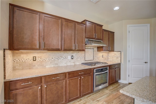 kitchen featuring tasteful backsplash, light stone countertops, under cabinet range hood, light wood-style flooring, and stainless steel appliances