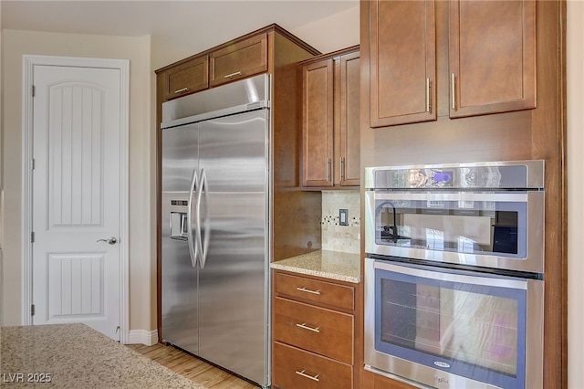 kitchen featuring light wood-type flooring, brown cabinets, tasteful backsplash, stainless steel appliances, and light stone countertops