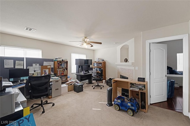 carpeted home office with a fireplace, a healthy amount of sunlight, visible vents, and ceiling fan