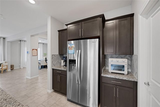 kitchen featuring stainless steel refrigerator with ice dispenser, tasteful backsplash, dark brown cabinetry, baseboards, and light stone countertops