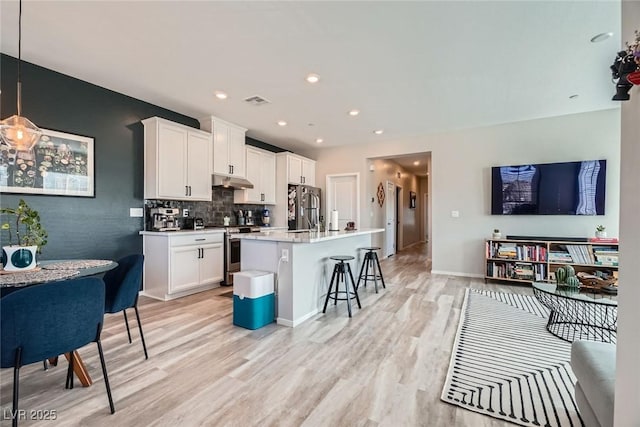 kitchen featuring light wood finished floors, under cabinet range hood, decorative backsplash, a kitchen breakfast bar, and stainless steel appliances