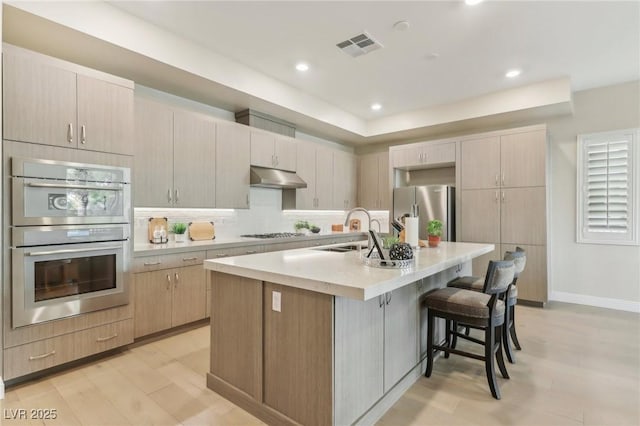 kitchen featuring a breakfast bar area, a center island with sink, a sink, stainless steel appliances, and under cabinet range hood