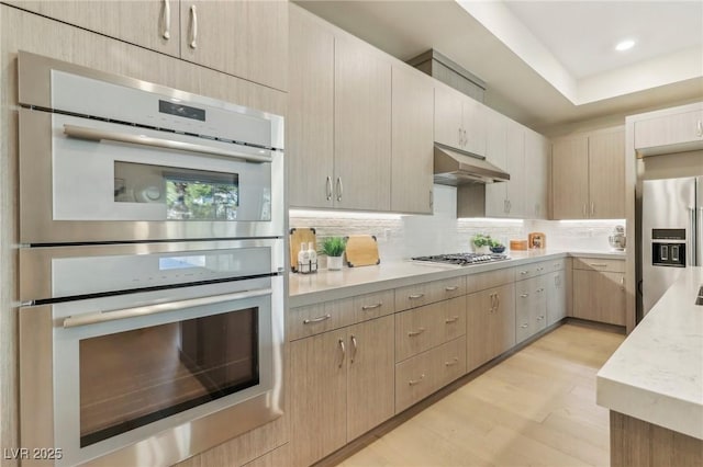 kitchen with light brown cabinetry, under cabinet range hood, tasteful backsplash, stainless steel appliances, and light wood-style floors