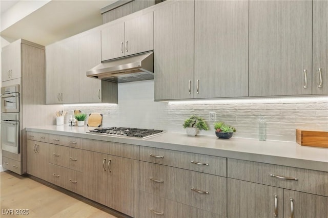 kitchen featuring under cabinet range hood, light countertops, decorative backsplash, light wood-style flooring, and stainless steel appliances