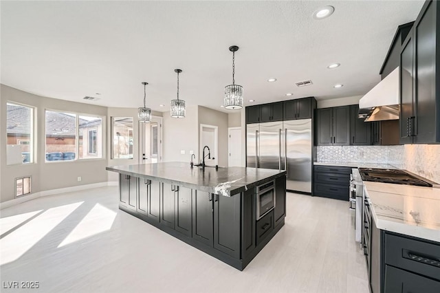 kitchen with visible vents, dark cabinetry, range hood, built in appliances, and a chandelier