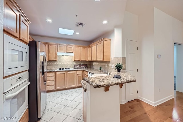 kitchen with visible vents, light brown cabinetry, under cabinet range hood, a peninsula, and white appliances