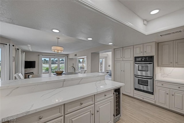 kitchen featuring visible vents, stainless steel double oven, wine cooler, pendant lighting, and light wood-type flooring