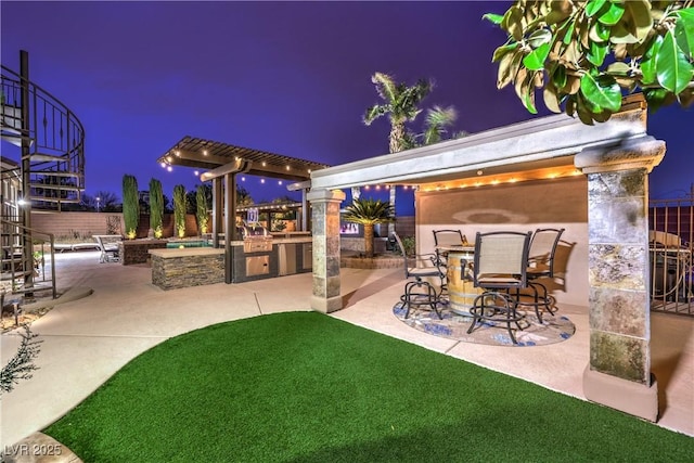 patio at night with outdoor dining space, exterior kitchen, fence, and a lawn