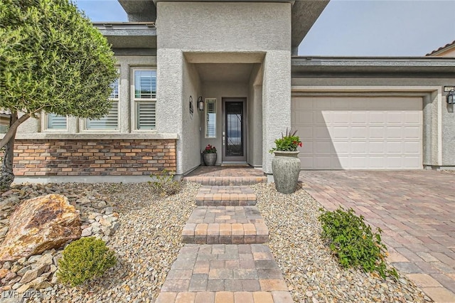 doorway to property featuring decorative driveway, an attached garage, brick siding, and stucco siding