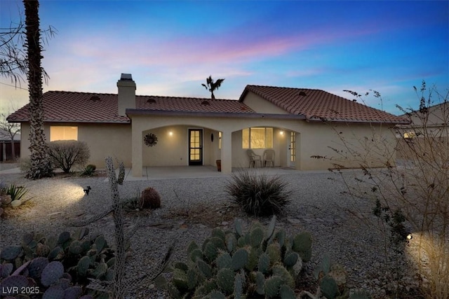 back of house at dusk featuring a chimney, a patio area, stucco siding, and a tile roof